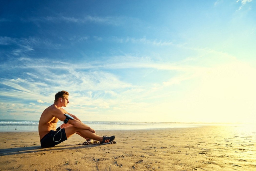 A man sitting on a sandy beach under a blue sky with clouds, surrounded by water and nature, enjoying a summer vacation.